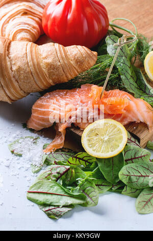 Slaced salted salmon with fresh salad leaves, lemon, sea salt and vegetables, served on wooden chopping board with croissant over light blue wooden ba Stock Photo