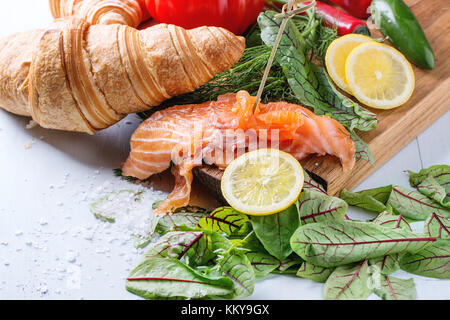 Slaced salted salmon with fresh salad leaves, lemon, sea salt and vegetables, served on wooden chopping board with croissant over light blue wooden ba Stock Photo
