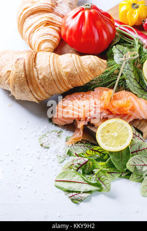 Slaced salted salmon with fresh salad leaves, lemon, sea salt and vegetables, served on wooden chopping board with croissant over light blue wooden ba Stock Photo