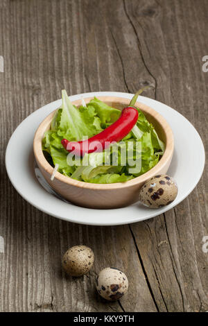 Wooden bowl of with green salad in white plate with red hot chili pepper and three quail's eggs on wooden table Stock Photo