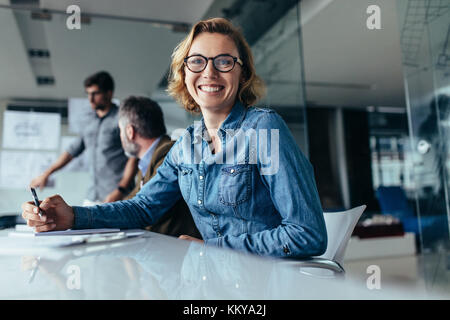 Caucasian businesswoman sitting in meeting with colleagues in background. Female designer sitting in board room looking at camera. Stock Photo