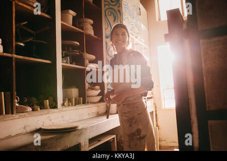 Smiling woman potter standing in her pottery workshop wearing an apron. Stock Photo