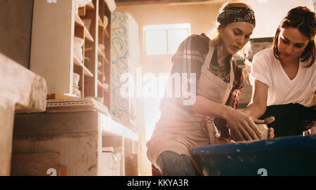 Female potter mentoring a woman in the craft of pottery. Young woman learning to mould clay on pottery wheel. Stock Photo