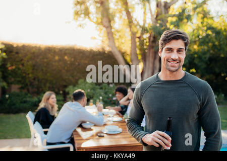 Handsome young man standing outdoors with friends sitting in background having food. Man with beer at outdoor party. Stock Photo