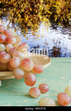 Hanging from wooden dish tassel of grape on wooden table against background of pond and hanging yellow leaves. Stock Photo