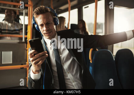 Focused young businessman wearing a suit listening to music and reading text messages while standing on a bus during his morning commute Stock Photo