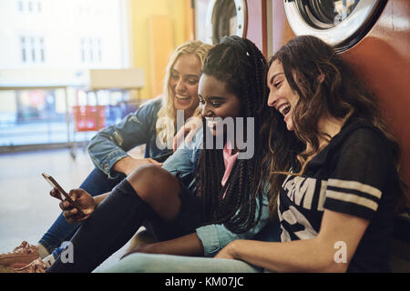 Group of smiling young girlfriends sitting on a laundromat floor using a cellphone together while doing laundry Stock Photo