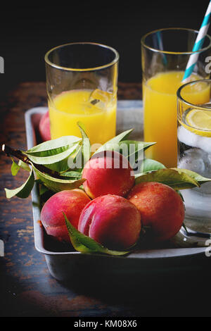 Peaches on branch with leaves and glasses with peach juice and limonade with ice cubes in aluminum tray over old metal table. Dark rustic style. Stock Photo