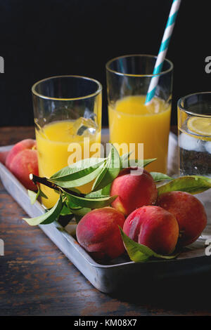 Peaches on branch with leaves and glasses with peach juice and limonade with ice cubes in aluminum tray over old metal table. Dark rustic style. Stock Photo