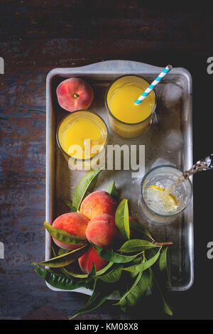 Peaches on branch with leaves and glasses with peach juice and limonade with ice cubes in aluminum tray over old metal table. Dark rustic style. Top v Stock Photo