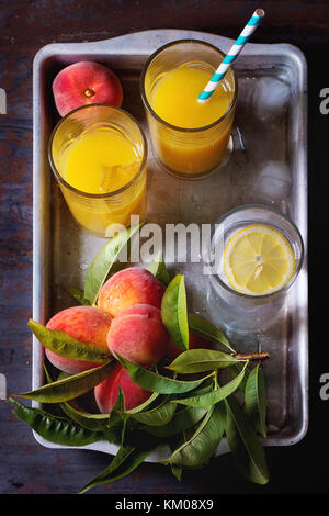 Peaches on branch with leaves and glasses with peach juice and limonade with ice cubes in aluminum tray over old metal table. Dark rustic style. Top v Stock Photo