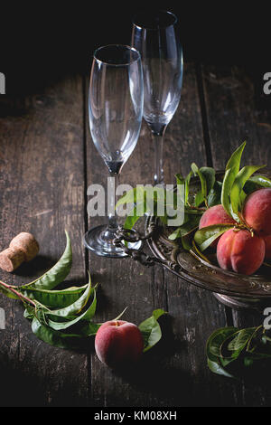 Peaches on branch with leaves in vintage plate and two empty glasses for champagne with cork over old wooden table. Dark rustic style. Stock Photo