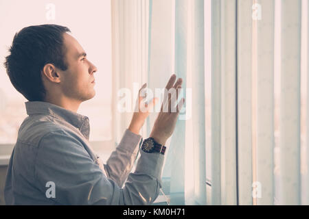 Young businessman standing in modern office, looking through blind, thinking. Vertical blinds light blue color on the big screen. Stock Photo