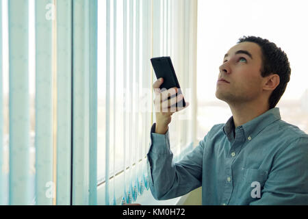 Young man using a smartphone in front of a window. Control vertical blinds with your phone. To open a window with a mobile app. Smart home. modern tec Stock Photo