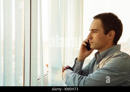 Businessman talking on the phone in office looking out the window. Vertical blinds light blue color on the big screen. Stock Photo