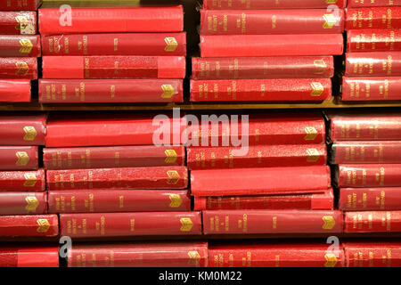 A stack or pile or red coloured hymn books and prayer books on a shelf ready for use in a church with gold letting on the spine. Singing religion. Stock Photo