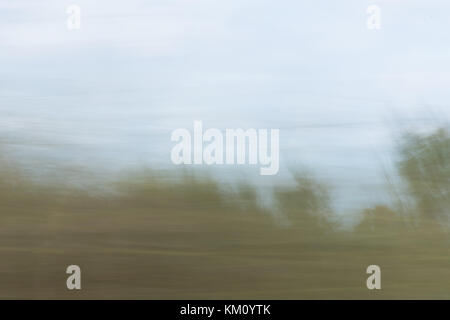 Blurred landscape photographed from a moving vehicle with tall green grass in the foreground and light blue sky above. Stock Photo