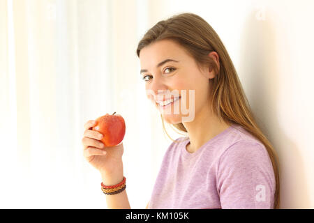 Portrait of a happy teen holding an apple and looking at you leaning on a wall in a house interior Stock Photo