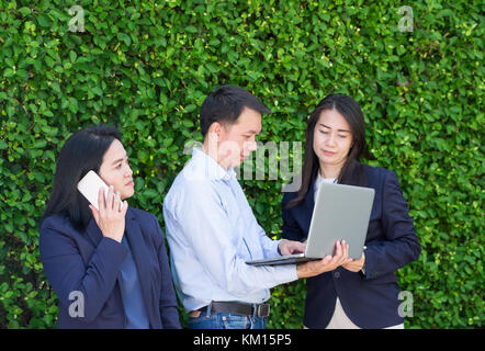 Businesswoman and businessman working outside office using laptop computer mobile phone and tablet device at green leaf wall,mobile office concept Stock Photo