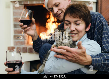 Man and woman playing with a kitten by the fireplace. They are holding glasses of wine. Stock Photo