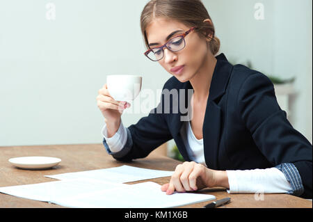 Pretty business woman reading data documents sitting at the table in office and drinking coffee Stock Photo