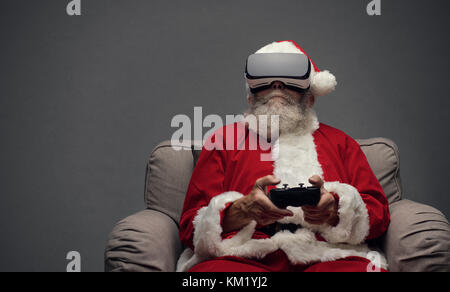 Santa Claus experiencing virtual reality at home and playing videogames using a controller, he is wearing a VR headset Stock Photo