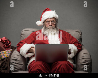 Santa claus relaxing at home and connecting with a laptop, he is chatting and social networking, Christmas time and technology concept Stock Photo
