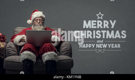 Santa claus relaxing at home and connecting with a laptop, he is chatting and social networking,Christmas card with wishes Stock Photo