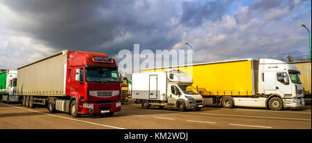 Colorful modern big semi-trucks and trailers of different makes and models stand in row on flat parking lot of truck stop in sunshine Stock Photo