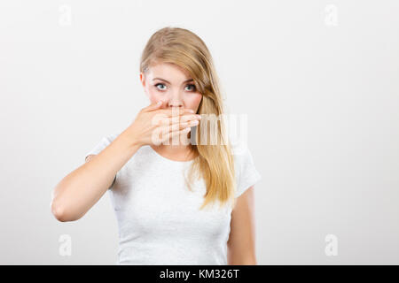 Saying bad things, shocking news concept. Ashamed young blonde woman having hand on her mouth. Studio shot on grey background. Stock Photo