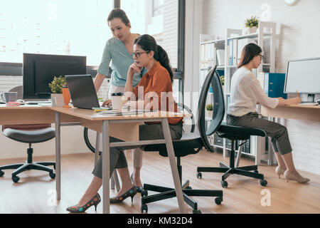 group of design team achieving best results. Two confident young women looking at laptop monitor while their colleagues working in the background. Stock Photo