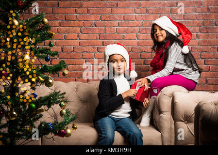 Indian kids celebrating christmas - cute little Indian kids playing, laughing and having fun with gift boxes on christmas, sitting on floor or sofa wi Stock Photo