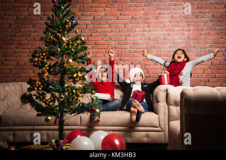 Indian kids celebrating christmas - cute little Indian kids playing, laughing and having fun with gift boxes on christmas, sitting on floor or sofa wi Stock Photo