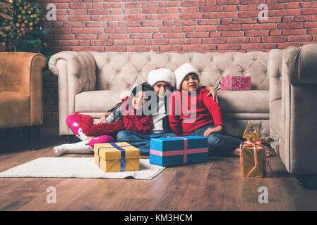 Indian kids celebrating christmas - cute little Indian kids playing, laughing and having fun with gift boxes on christmas, sitting on floor or sofa wi Stock Photo