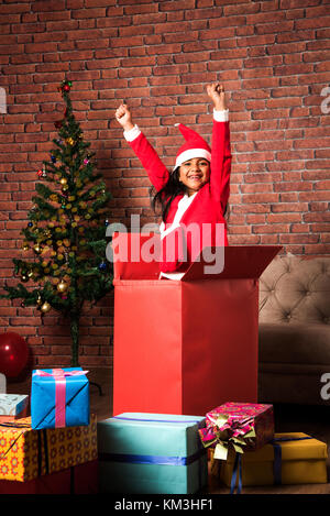 surprised Little Indian girl standing inside or coming out of big gift box in santa clause attire on christmas evening with decorated tree in the back Stock Photo