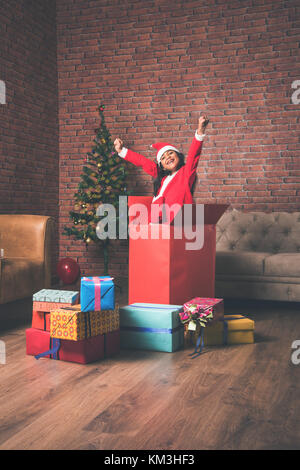 surprised Little Indian girl standing inside or coming out of big gift box in santa clause attire on christmas evening with decorated tree in the back Stock Photo