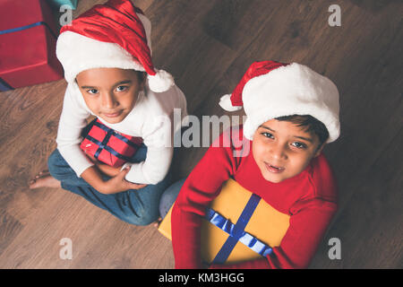 Indian kids celebrating christmas - cute little Indian kids playing, laughing and having fun with gift boxes on christmas, sitting on floor or sofa wi Stock Photo