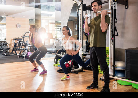 Young determined fit woman doing squats exercise while holding on the back of her neck a heavy sandbag, during group circuit functional training at th Stock Photo