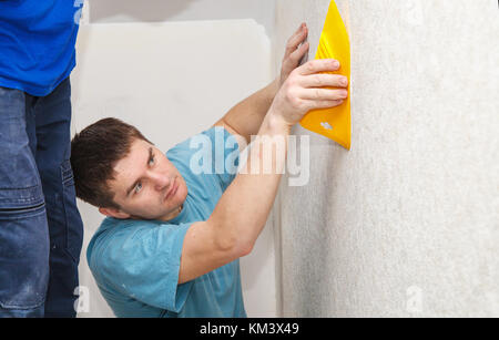 young worker with spatula smoothing wallpaper in his house Stock Photo