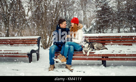 Christmas portrait of the loving couple sitting on the bench while their siberian husky lying near them during the snowfall in the forest. Stock Photo