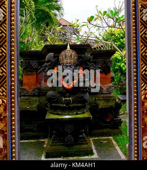 Vertical shot of statue of elephant-headed Hindu god Ganesha (Ganesh) with marigold garland in leafy temple garden, Ubud, Bali framed by ornate border Stock Photo