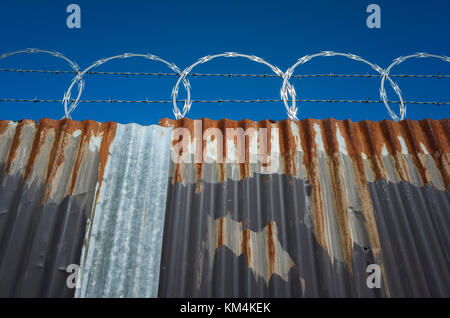 Low angle view of worn corrugated metal fence, razor wire above. Stock Photo