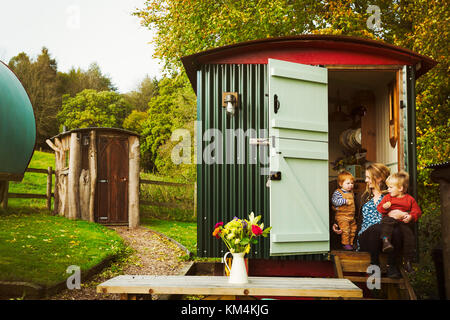 A shepherd's hut with open door beside a path to a small rustic shed, and a woman with two small children seated on the step. Stock Photo