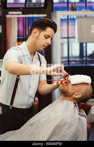 A customer sitting in the barber's chair, with a hot towel on his face, and a barber trimming his beard. Stock Photo