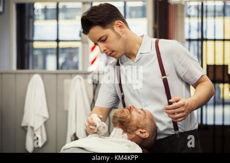 A customer sitting in the barber's chair, having a wet shave. A man using a shaving brush to lather his chin and neck. Stock Photo