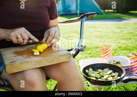 A person sitting beside a camp stove preparing yellow zucchini, courgettes for the frying pan. Stock Photo