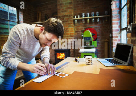 A craftsman using an awl and a ruler to measure and mark a small piece of leather.  Workbench with a mug, hand tools and a laptop. Stock Photo