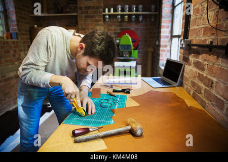 A craftsman using a rotary cutter wheel on a cutting mat on a small piece of brown leather. Stock Photo