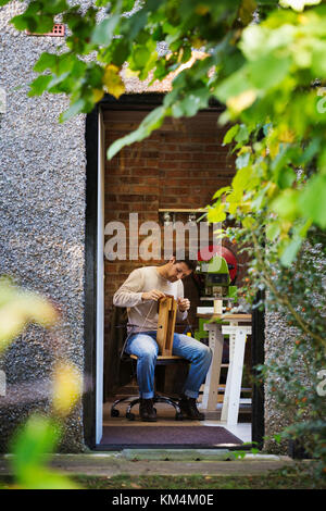 View through an open door of a craftsman leatherworker seated in his workshop, stitching together a small leather object. Stock Photo