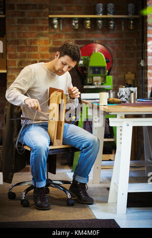 A craftsman leatherworker seated in his workshop, stitching together a small leather object. Stock Photo